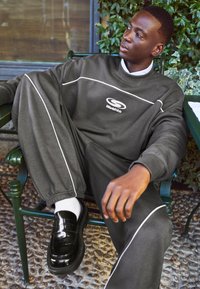 Siv športni pulover in hlače z belimi detajli, logo na prsih, črne sijoče čevlje, sede na zeleni stol s foliagom v ozadju.