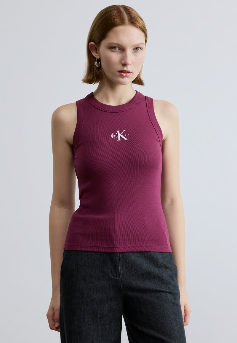 Lila bordázott ujjatlan felső magas nyakkal és egy kis fehér Calvin Klein logóval a mellkason. A modell sötét farmernadrágot visel.