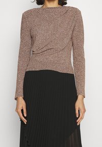 Top a maniche lunghe in un tessuto metallico color rosa dorato a trama con una gonna nera plissettata. Il corpetto presenta un dettaglio drappeggiato in vita.