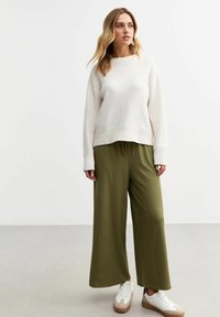 Pull en tricot beige clair avec ourlet à côtes, associ�é à un pantalon large plissé vert olive foncé et des baskets blanches avec des accents marron.