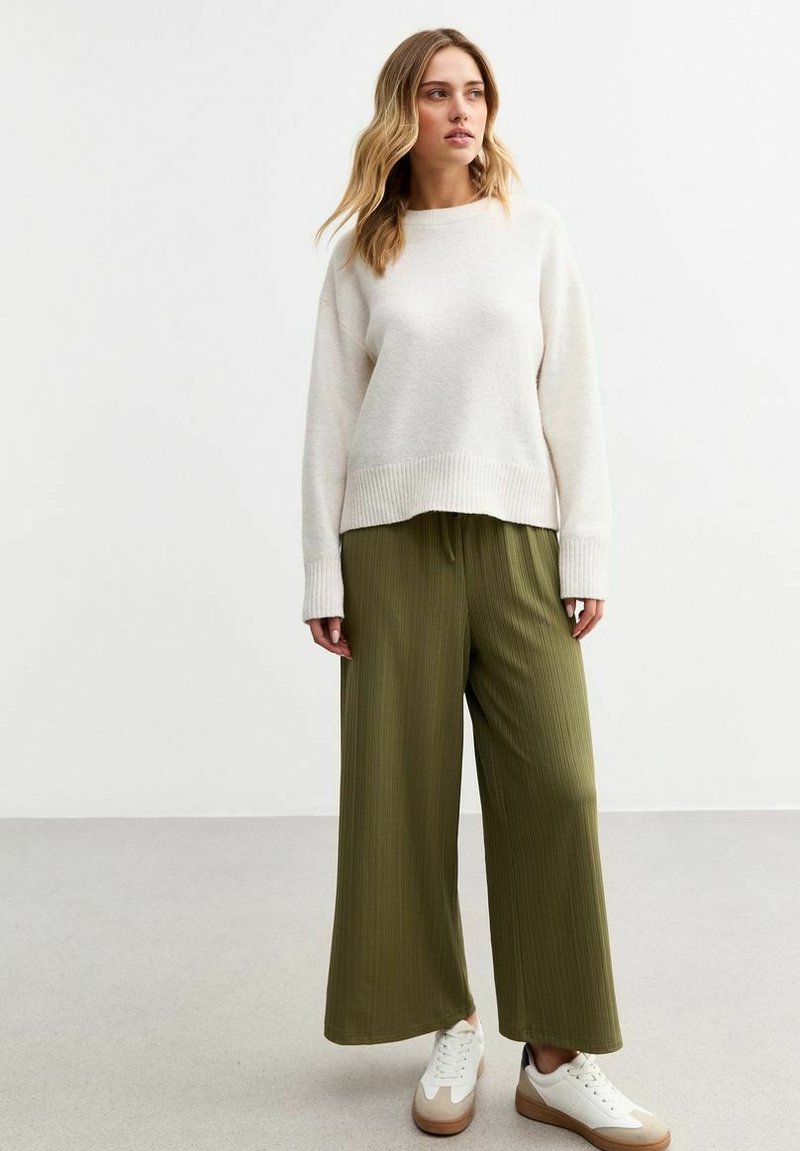 Pull en tricot beige clair avec ourlet à côtes, associé à un pantalon large plissé vert olive foncé et des baskets blanches avec des accents marron.