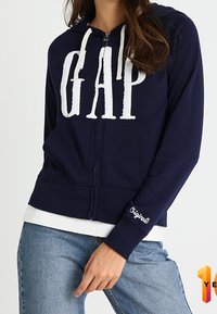Marineblauwe zip-up hoodie met een opvallend wit "GAP" logo. Voorzien van koordjes, voorzakken en een geribbelde zoom. Gdragen over een witte top.
