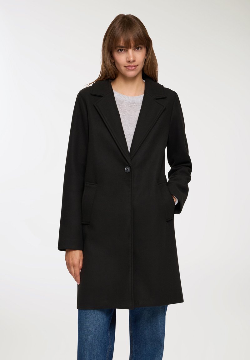 Cappotto di lana nera con collo a scollatura, un solo bottone, due tasche frontali e taglio dritto. Indossato sopra una maglia grigia chiara con jeans blu.