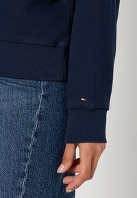 Felpa navy con polsini e orlo a coste, con un piccolo etichetta del logo del marchio in rosso, bianco e blu, abbinata a jeans blu.