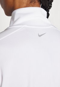 Valge kõrge kaelusega spordipoli, millel on sile tekstuur, sisaldab vä Small halli Nike logo �ülemisel seljal ja peeneid õmblusdetail. 
