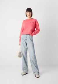 Sudadera corta rosa con un logotipo bordado, combinada con vaqueros de pierna ancha en azul claro y zapatos en blanco y negro, sosteniendo un bolso blanco.