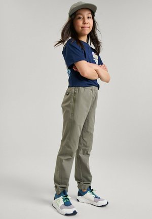 Pantaloni verdi leggeri a gamba affusolata con polsini elastici, abbinati a una maglietta blu navy e un berretto grigio, completati da sneakers bianche e blu.