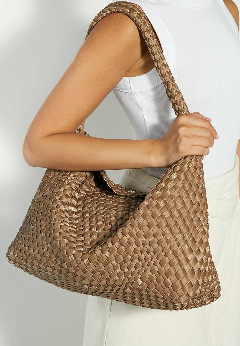 Femme tenant un grand sac d'épaule en osier marron avec une bandoulière épaisse, portant un haut blanc sans manches et une jupe de couleur claire.