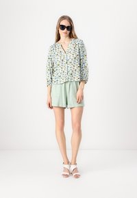 Floral print blouse with balloon sleeves, button front; light green shorts with two pockets; modeled with black sunglasses and white sandals.