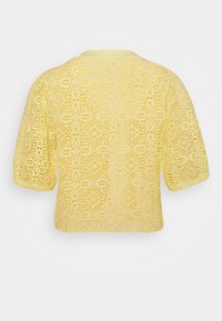 Blouse jaune à manches courtes en dentelle avec motif floral et encolure ronde, vue de dos sur un fond uni.