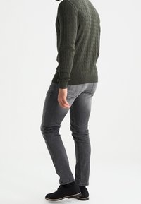 Tmavě zelený svetr s pleteným vzorem, šedé slim-fit džíny a černé boty, foceno zezadu. Na svetru jsou viditelné texturované detaily.