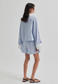 Camicia a righe blu e bianche, ampia e comoda, con polsini bianchi a contrasto, abbinata a pantaloni corti coordinati. Il modello indossa sandali neri e occhiali da sole.