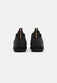 Zapatillas deportivas negras con un acabado texturizado, que presentan un acento dorado y un logo en el talón, así como tiradores para facilitar su uso.