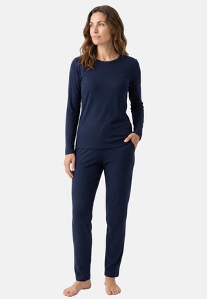 Woman standing barefoot wearing a navy blue long-sleeve top and matching pants, looking to the side with one hand in pocket.