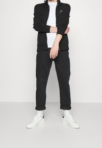 Mies, joka on pukeutunut mustaan vetoketjulliseen takkiin, valkoiseen t-paitaan, mustiin lahkeisiin housuihin, valkoisiin sukkien ja valkoisiin lenkkikenkiin, seisoo yksinkertaista valkoista taustaa vasten.