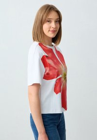 Jonge vrouw met steil schouderlang haar, gekleed in een witte t-shirt met een grote rode bloemenprint en blauwe spijkerbroek, staand in profiel.
