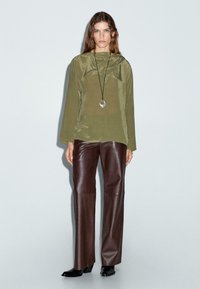 Haut à manches longues vert olive avec capuche et coupe décontractée, associé à un pantalon en cuir brun et des chaussures noires à bout pointu, agrémenté d'un collier avec pendentif.