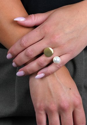 Zwei Ringe an einer Hand: Der eine ist eine polierte Goldscheibe, der andere eine runde weiße Perle, beide vor leicht manikürten Fingernägeln platziert.