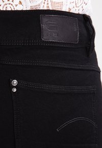 Mustat denimit, joissa on nahkaleima kohokuvioidulla logolla, hopeiset metalliosat ja erottuvat tikkaukset taskuissa.