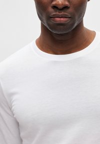 Balta medvilnės marškinėliai su apvalia iškirpte ir lygiu paviršiumi. Artimas vaizdas, akcentuojantis audinį ir iškirptės detalę.