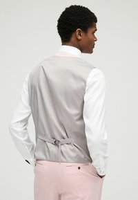 Gilet grigio in satin con una fascia sul retro, abbinato a una camicia bianca. I pantaloni sono di un rosa chiaro con due tasche posteriori e dettagli a bottoni.