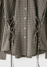 Chemise à carreaux marron et blanc avec des boutons au centre et des lacets de chaque côté près de la taille.