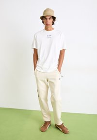 Hvid bomulds T-shirt med rund hals, beige bøttehat, cremefarvede joggere med sidelommer og lysebrune sneakers på et grønt gulv.