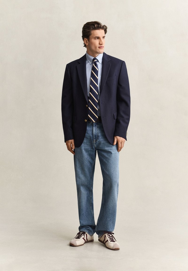 Mand iført navy blazer, lyseblå skjorte med stribet slips, blå jeans og hvide sneakers står foran en ensfarvet baggrund.