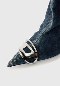 Accesorio de denim con una hebilla metálica plateada que tiene un recorte rectangular. El denim presenta una textura con costuras visibles y un tono azul clásico.