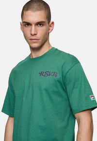 Groene katoenen T-shirt met een paarse "R.S.V.P."-logo op de borst en een klein Fila-logo op de mouw. Korte mouwen en een klassieke pasvorm.