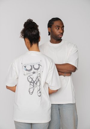 Two people in white t-shirts; one faces forward with arms crossed, the other shows a graphic cassette tape and tangled tape design on back.