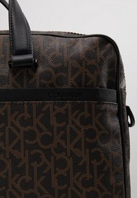 Sac Calvin Klein en matériau texturé marron avec un motif de logo répétitif. Poignées et garniture en cuir noir, forme rectangulaire avec fermeture éclair.