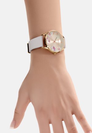 Montre dorée avec un cadran rond orné de motifs floraux, un bracelet en silicone blanc et une boucle classique. L'affichage présente des chiffres et des marqueurs.