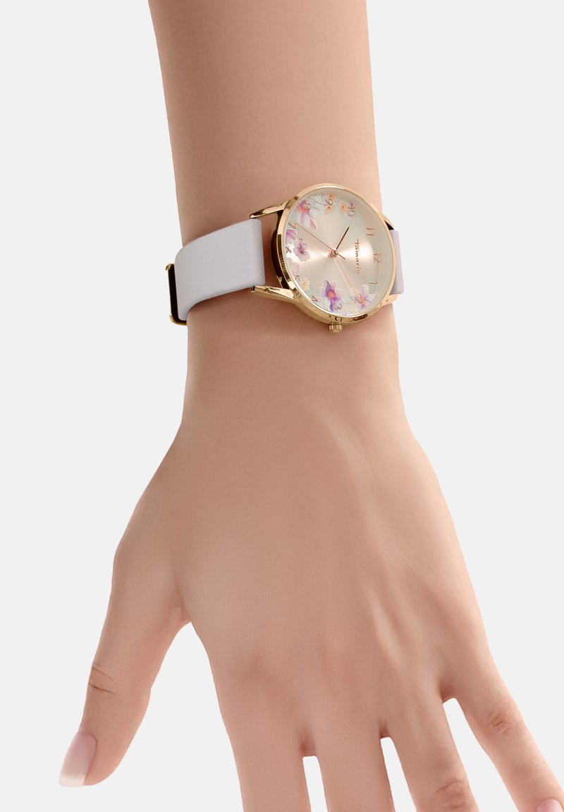 Goldfarbige Uhr mit rundem Ziffernblatt, das florale Muster zeigt, einem weißen Silikonarmband und einem klassischen Verschluss. Das Display zeigt Zahlen und Markierungen.