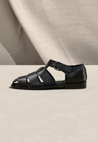 Sandale de pêcheur pour homme en cuir noir avec plusieurs brides, boucle à la cheville et semelle plate, présentée sur une surface neutre.