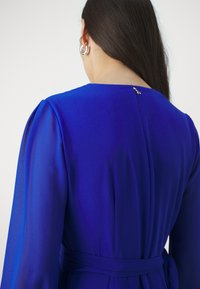 Plava haljina s dugim rukavima, izrađena od glatkog materijala. Ima suptilni zatvarač na leđima i pojas s vezanjem u struku, s elegantnim siluetom.