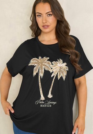 Frau mit langen braunen Haaren, die ein schwarzes T-Shirt mit zwei Pailletten-Palmen und dem Schriftzug "Palm Springs Beach Club" trägt.