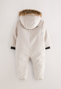 Smėlio spalvos izoliuotas kombinezonas su kailinėmis apykaklės krašteliais, juodos spalvos akcentais ant rankogalių, tampriai prilaikoma apačia ir siuvinėtu prekės ženklo ženklinimu nugaroje.