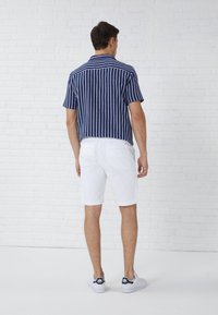 Chemise à rayures bleues et blanches à manches courtes associée à un short blanc. Le mannequin porte des baskets blanches. L'arrière-plan présente un mur en briques blanches.
