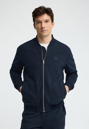 Man draagt marineblauwe jas met rits en bijpassende broek, staand met handen in de jaszakken tegen een effen achtergrond.