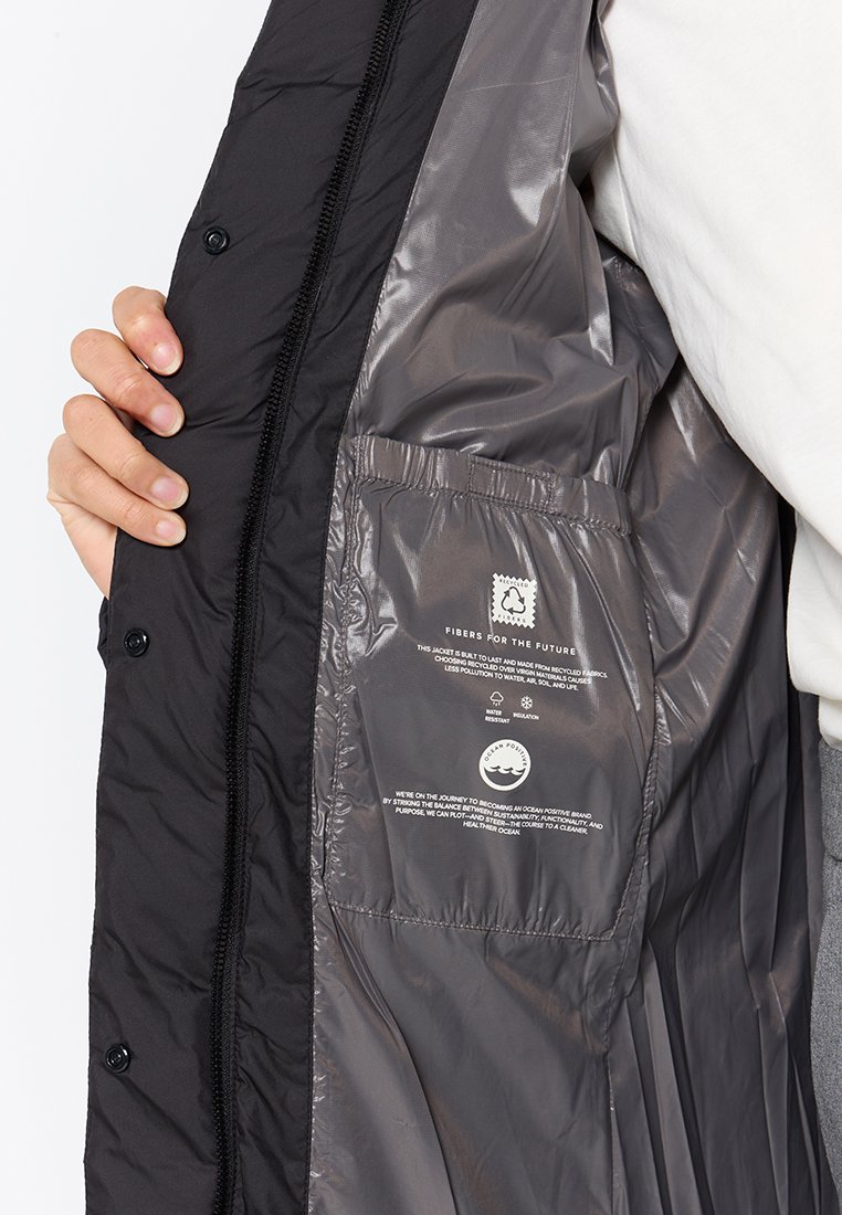 Una mano tiene aperto un giubbino nero che rivela una fodera interna grigia con tasca e un'etichetta di sostenibilità riguardante le fibre riciclate e la cura degli oceani.