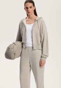 Světle šedá zkrácená mikina z měkkého materiálu s klokankovými kapsami, v kombinaci s odpovídajícími tepláky. Drží béžovou polstrovanou tašku.