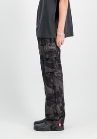 Svart camouflage cargo-byxor med flera fickor, förstärkt sömnad och en broderad "ALPHA"-logotyp på baksidan. Matchas med svarta skor.