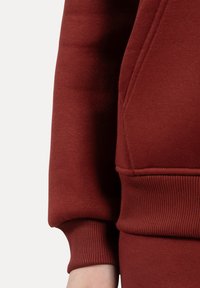 Felpa bordeaux con polsini e orlo a costine. Tessuto spesso e liscio con una tasca frontale e dettagli cuciti. Texture morbida evidenziata lungo i bordi.