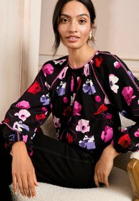 Blusa nera a fiori con motivi rosa, viola e bianchi. Presenta maniche a sbuffo e dettagli a scallop lungo il collo e i polsini.