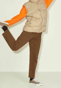 Beige pufferväst över en orange långärmad tröja, tillsammans med bruna byxor och bruna sneakers. Bakgrunden är i ljusa färger.