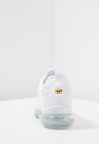 White sneaker with a mesh and synthetic upper, transparent air cushioning sole, and a yellow and red logo badge on the heel.