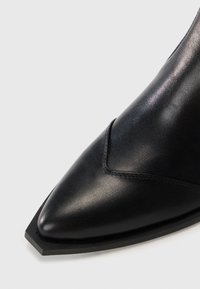 Botă din piele neagră până la gleznă, cu vârf ascuțit, textură netedă și toc geometric. Prezintă detalii subtile de cusătură pe partea superioară.