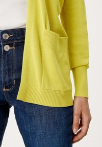 Gul stickad cardigan med ribbade ärmar och nederkant, med en framfick, i kombination med mörkblå jeans. Närbild.