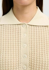 Gilet en tricot beige avec un motif texturé, un col, et six boutons de fermeture avec des boutons ronds de couleur claire.